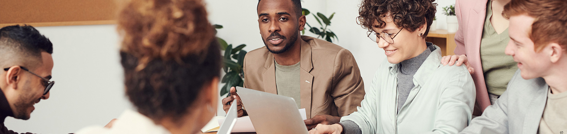 Eine Gruppe von Personen sitzt an einem Tisch und arbeitet zusammen. Auf dem Tisch liegen ein Laptop und verschiedene Unterlagen. Im Hintergrund sind Pflanzen und eine Pinnwand zu sehen.